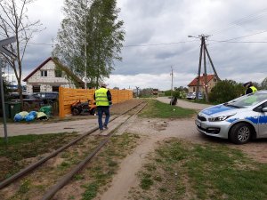 Miejsce zdarzenia drogowego, w centrum zdjęcia technik kryminalistyki zabezpieczający ślady na torach wąskotorówki, po prawej stronie oznakowany radiowóz