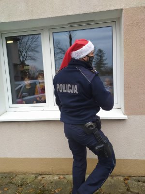 Policjant w czapce Mikołaja macha do dzieci przed przedszkolem