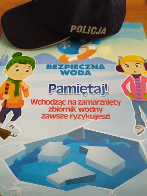 plakat mówiący o tym, że wchodzenie na zamarznięte zbiorniki jest niebezpieczne, na plakacie leży czapka z napisem policja